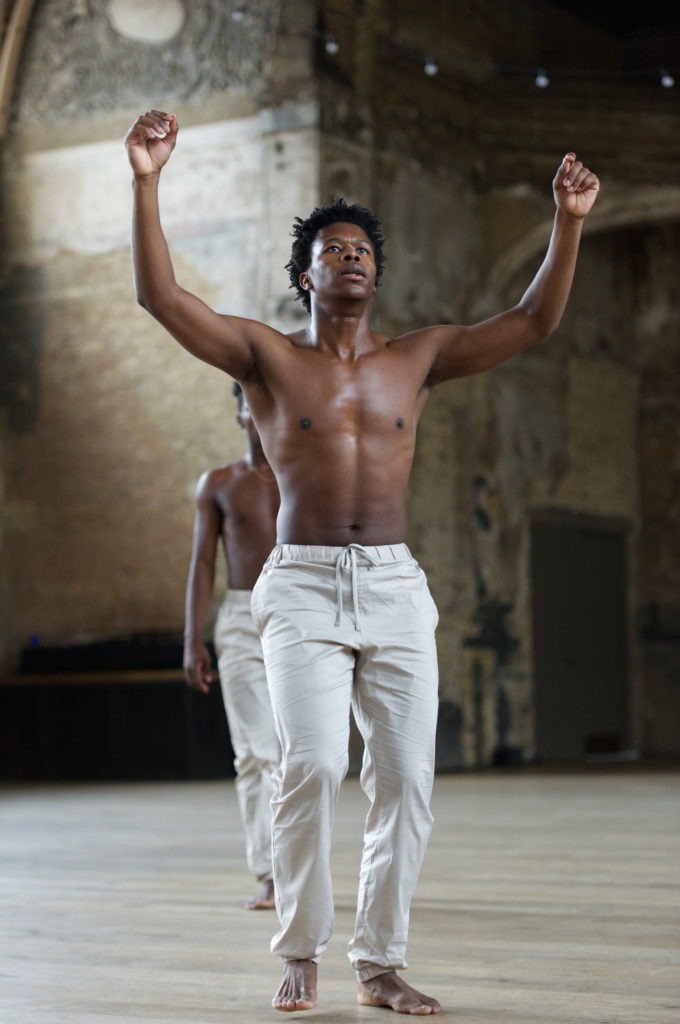 In a large, grungy space, Mthuthuzeli November opens his arms to either side of his head, elbows bent. He is bare-chested and wears white sweatpants. His gaze is lifted above the camera. He mostly hides a similarly outfitted dancer, walking up behind him.
