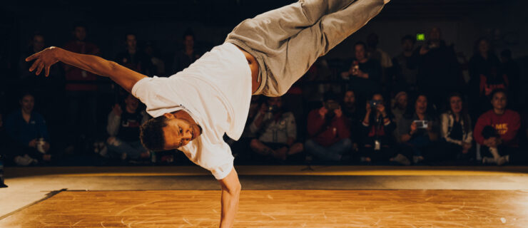 Morris "Bboy Morris" Isby balances on one arm, his legs both extended to 2 o'clock while his head is at 8 o'clock. He wears a white t-shirt, grey sweats, and well worn black Converse. Many members of the crowd watching at the side of a wooden dance floor have expressions of shock and delight.