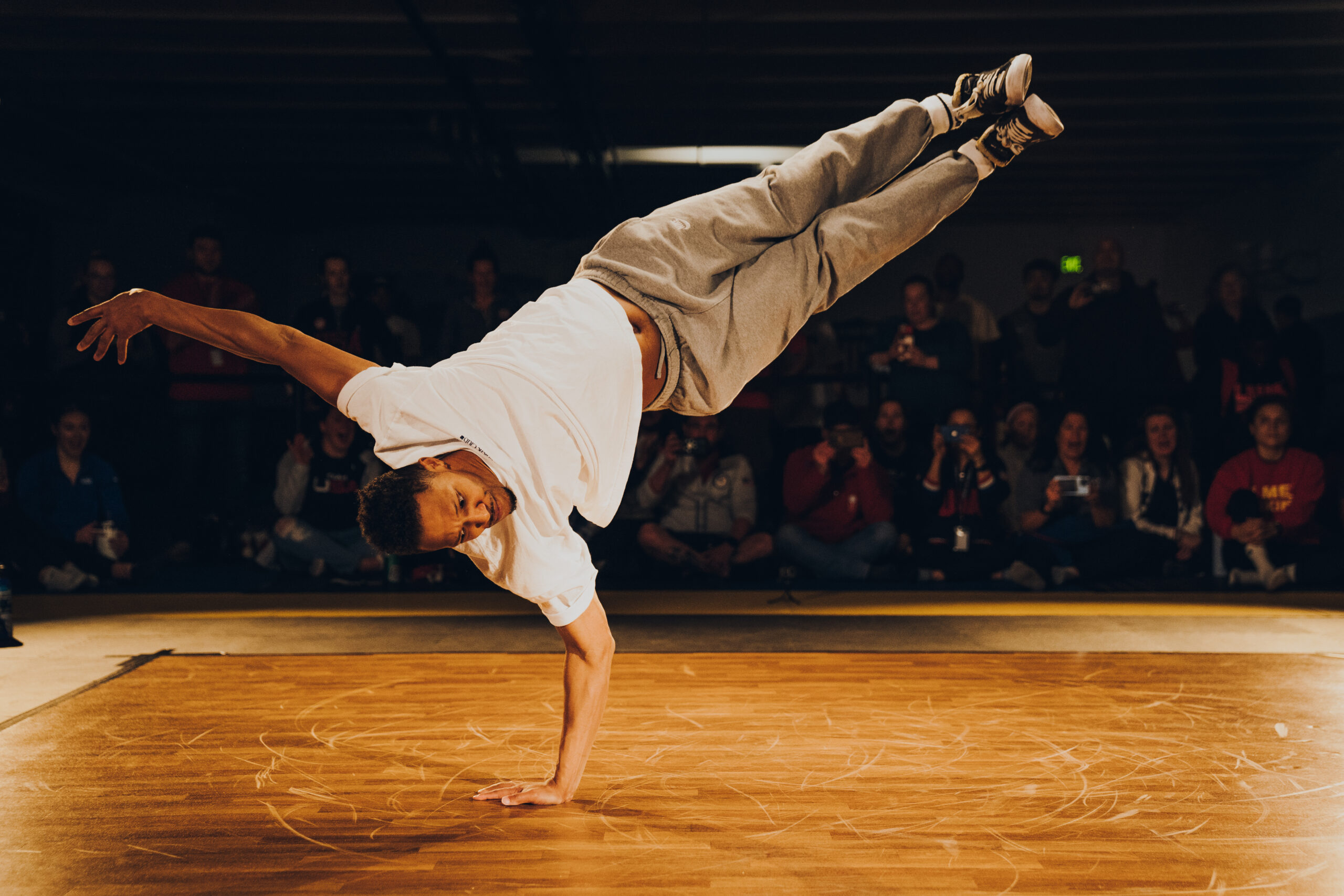 Morris "Bboy Morris" Isby balances on one arm, his legs both extended to 2 o'clock while his head is at 8 o'clock. He wears a white t-shirt, grey sweats, and well worn black Converse. Many members of the crowd watching at the side of a wooden dance floor have expressions of shock and delight.
