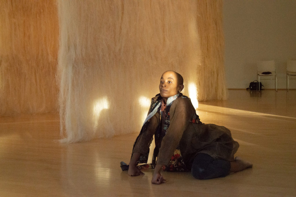 Jasmine Hearn kneels on the hardwood floor of a large room, their face looking forward in a stream of light