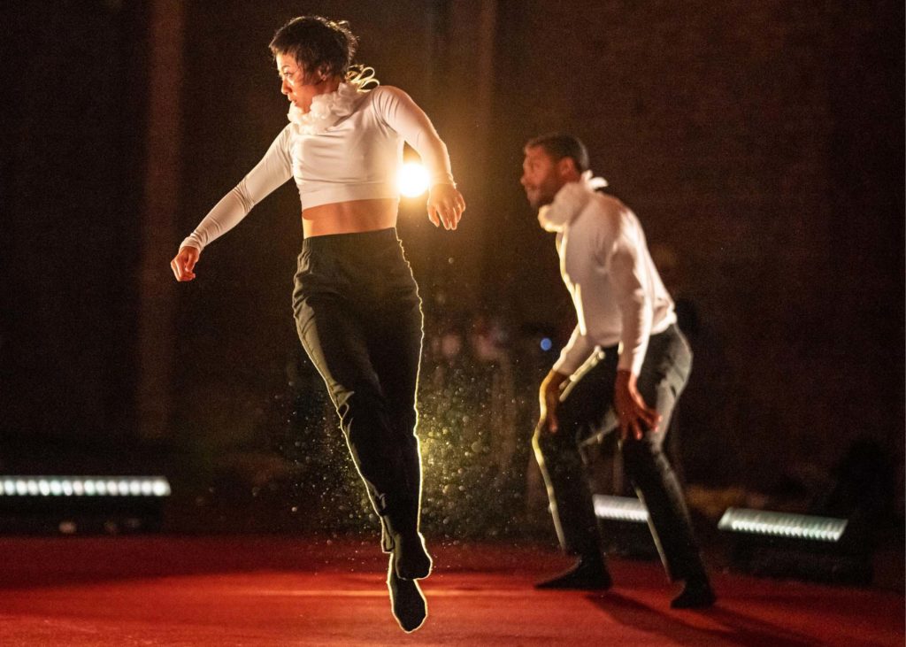 Two dancers in black pants, long sleeved white shirts, and white frilly collars are seen on a red carpet. In the foreground, a dancer jumps, legs extended below, arms lifting to her sides, face turned to the front of the performing space. In the background, the second stands in second position plié, hands splayed against his knees as he leans slightly forward.