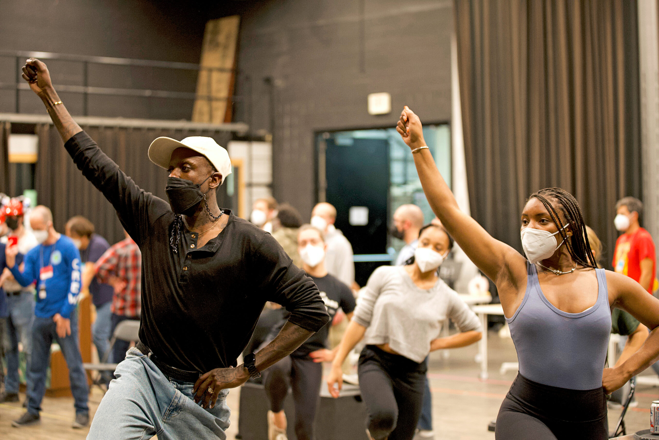 Raja Feather Kelly demonstrates in a rehearsal room, one hand upraised as though it holds a lit torch, same-side hip hitching upward. He wears a white cap, a black face mask, a black t-shirt, and sweats.