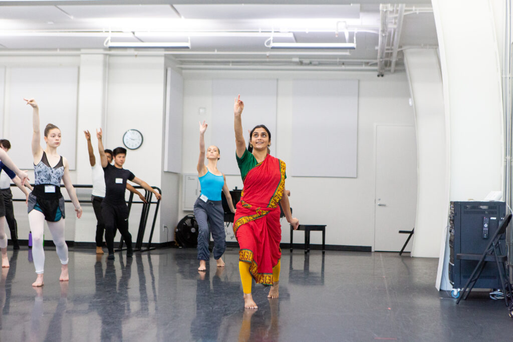 a female instructor in classical Indian attire leading students in a dance studio