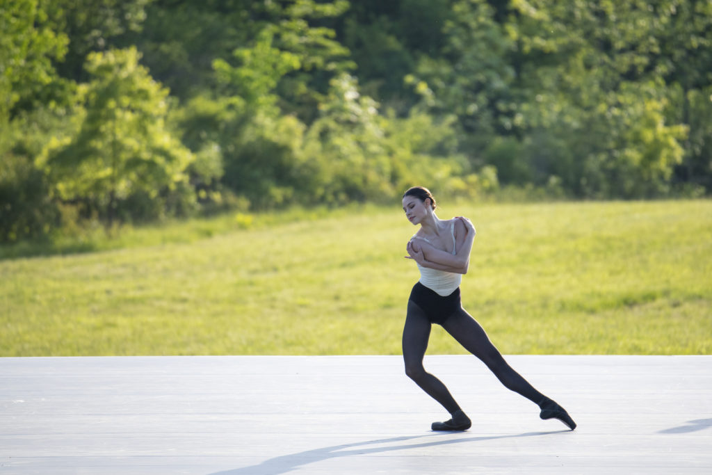 In black tights and pointe shoes and an off-white leotard, Sierra Armstrong poses in a tendu side, standing leg in plié. Her hands hug her upper arms as she gazes serenely away from her working leg. Grass and trees are visible beyond the marley on which she dances.