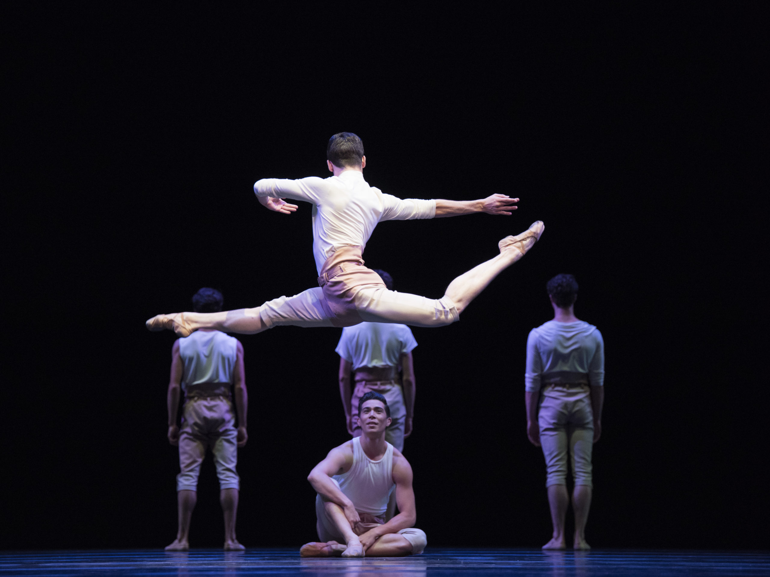 In the foreground, a male dancer leaps with his back leg in attitude, torso twisting to face upstage, arms in opposition to legs. Another dancer sits on the floor, looking up at him. In the shadowy upstage, three dancers stand with their backs to the audience. All wear white shirts, khaki shorts, and ballet slippers.