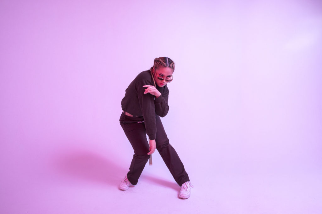 Sienna Lalau poses against a pink backdrop. She throws an intense look over her sunglasses as she hunches forward, one arm dangling in front of her purposefully turned in knees. She wears all black, except for a pair of worn white sneakers.