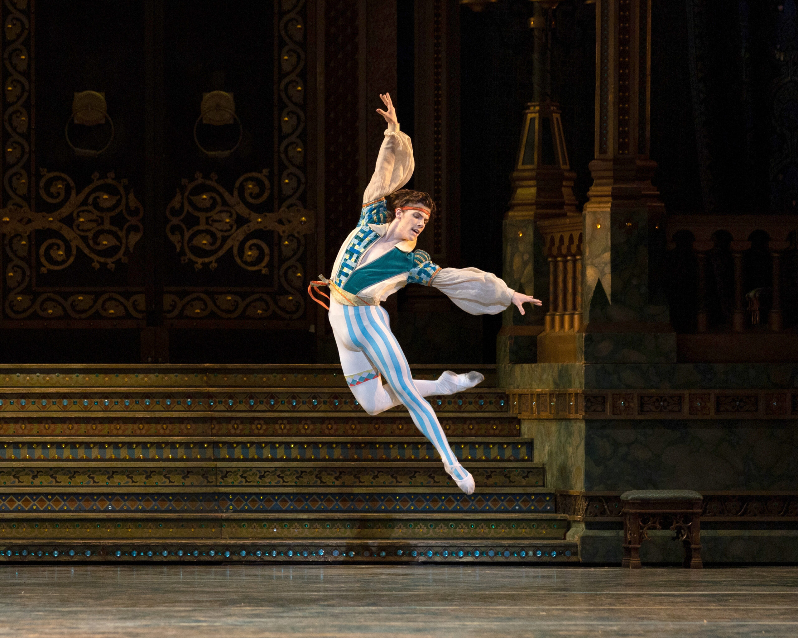 Jake Roxander is captured midair, arching back as he jumps in an overcrossed retiré back position. His costume features blue and white stripes down one leg and teal patterns on his puffy-sleeved tunic. The set behind him evokes an opulent ballroom.