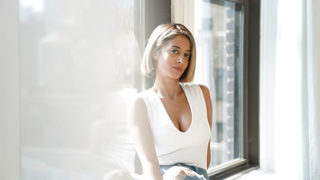 Iranian choreographer Tina Bararian leans against a window wearing a white v-neck shirt and jeans.