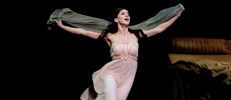 Mayara Magri is captured mid-run, arms outstretched to the sides as the cloak she holds in her hands billows behind her. Her costume is a pale peach dress over pink tights, reminiscent of a nightgown. Her hair is loose around her shoulders, a look of mingled despair, fear, and hope on her face.