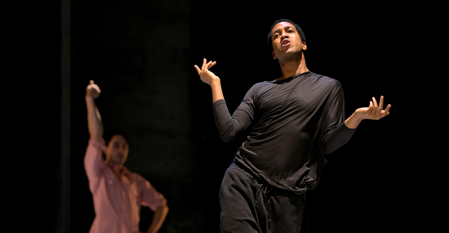Trajal Harrell, a slender Black man, leans into one hip as he steps forward. His elbows hug to his ribs as he raises his hands and chin. He is dressed in all black, while another dancer in the background wears a pink button-down as they mime holding something overhead with one hand.