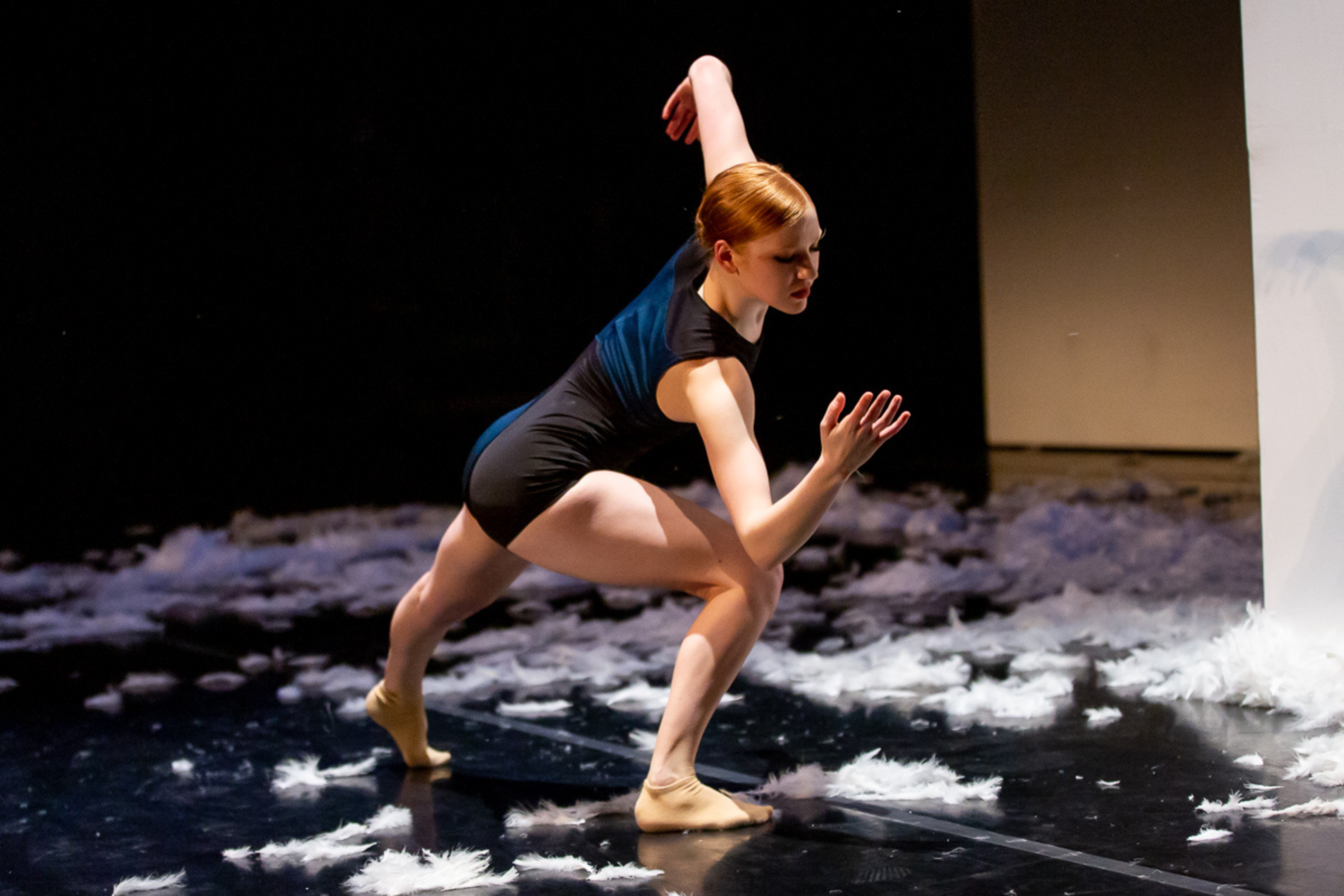 Lizzie Trip lunges to the side, an elbow bending to touch her bent knee. We see her from the wings, her back to the audience and face to the audience. White fluff litters the floor of the stage all around. Lizzie is barefoot, wearing a tight black and blue biketard, red hair neatly slicked back from her face.