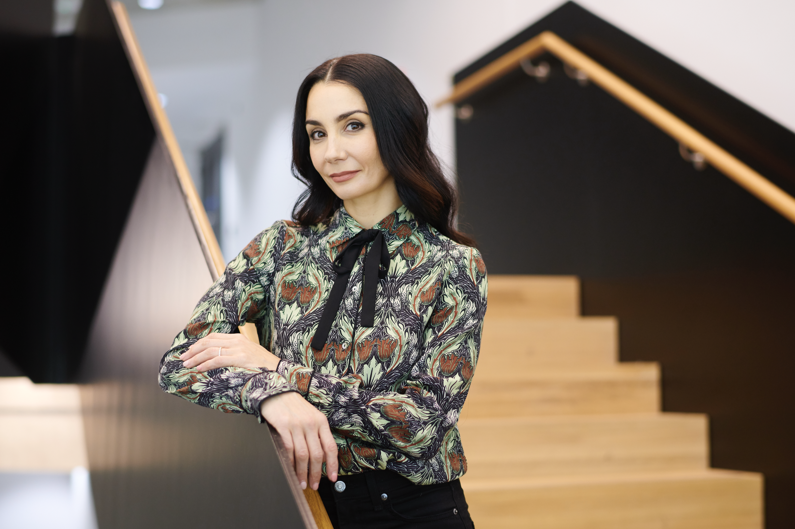 Tamara Rojo stands on a staircase, her arms resting on the railing, looking at the camera in a business casual outfit