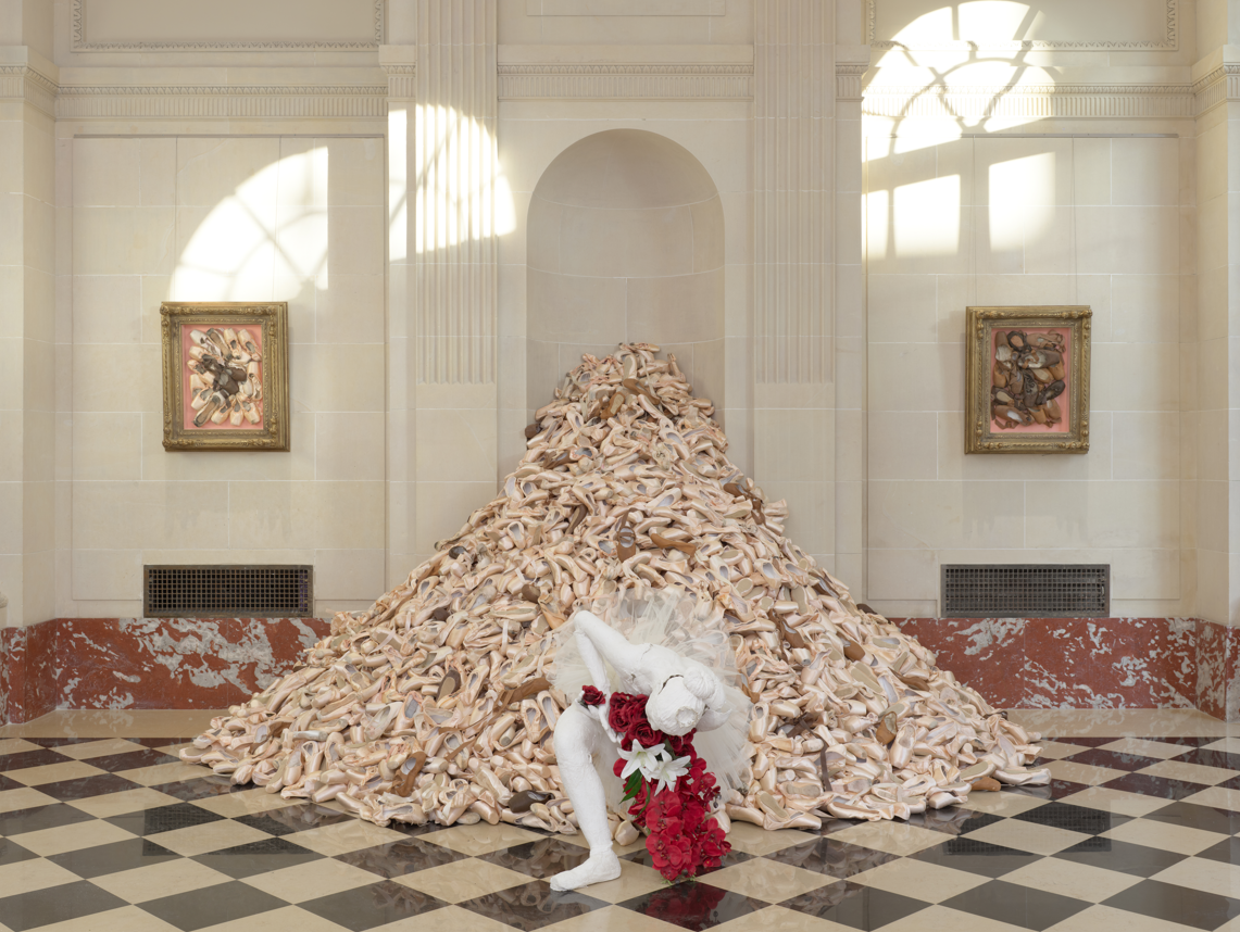 a large pile of pointe shoes with a bowing female ballerina mannequin in front of it