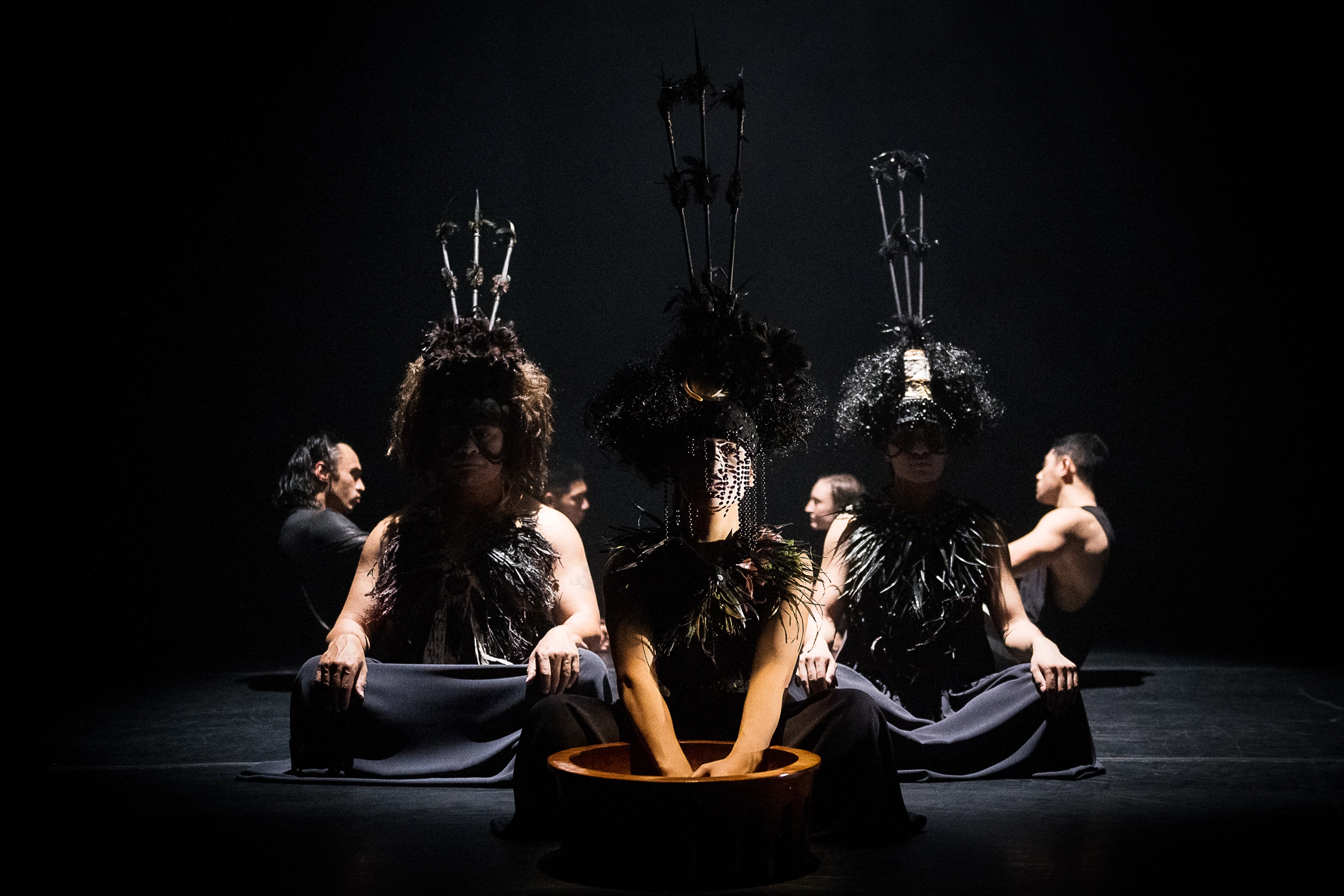 In the foreground, three dancers sit facing downstage, legs folded and crossed. The dancer at the center reaches hands into a deep brown bowl. All three wear collars that evoke feathers and headpieces with a trio of sticks pointing straight up. The downstage dancer has dark beads dangling in front of their face; the other two wear masks that cover their foreheads and around their eyes.