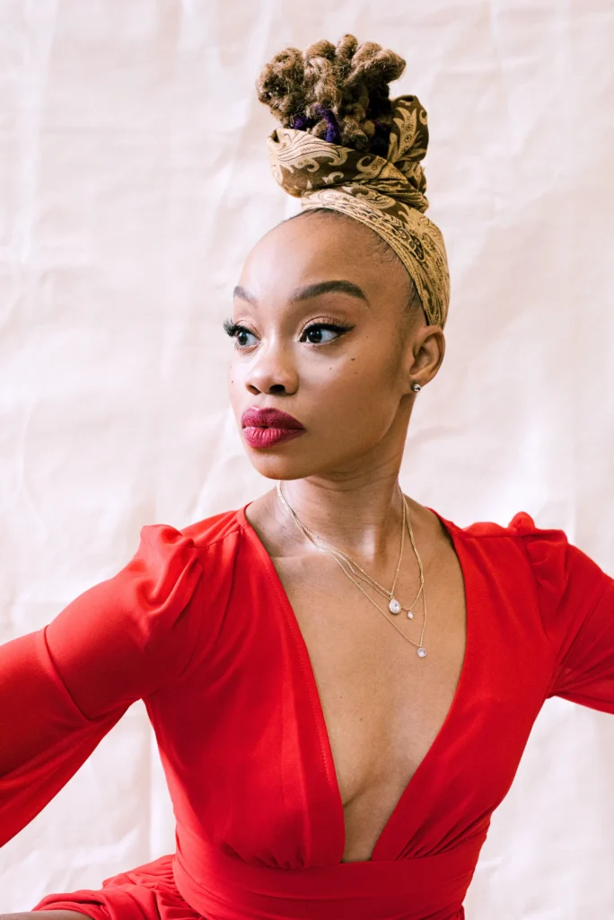 Camille A. Brown looks over her right shoulder. She wears a red blouse with a plunging neckline; her lips are painted the same color. A headscarf with a gold filigree pattern is wrapped around her scalp and some of the hair piled atop her head.