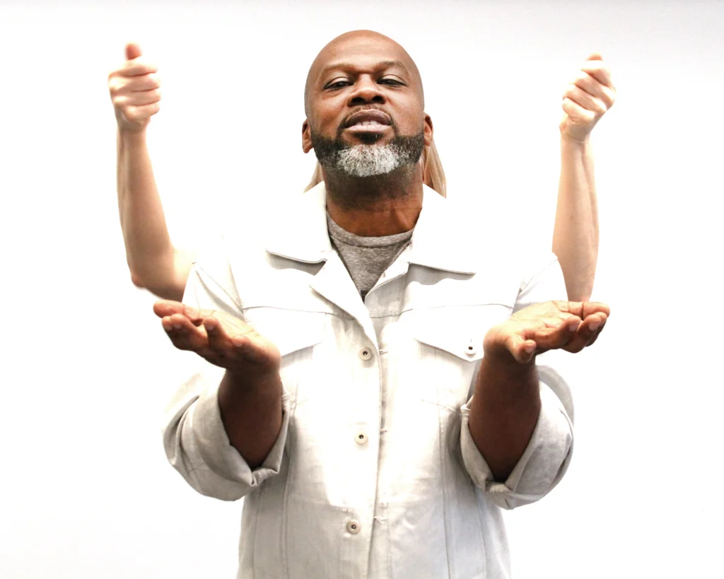 Marc Bamuthi Joseph stands against a white backdrop, palms upraised in offering as his arms bend at the elbow. Wendy Whelan is almost invisible behind him, save for her paler arms rising up from behind his shoulders, hands in loose fists.