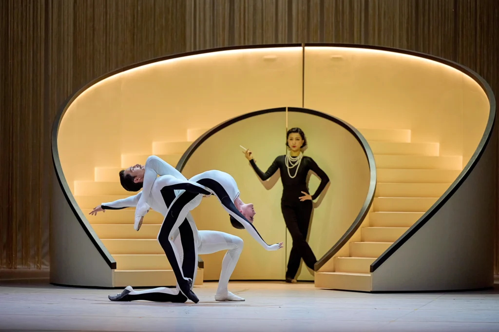 Upstage, a woman in head to toe black and draped pearl necklaces stands with a hand on her hip and a cigarette in the other, leaning against the base of a set of circular stairs. Her gaze is focused on two dancers downstage, each wearing white unitards with black side stripes. The dancer en pointe arches back toward the floor, her extended leg draped over her partner's shoulder. He kneels facing her and supports her at the waist, head tipped back to mirror her arch.
