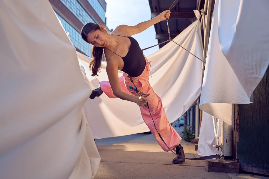Amidst draped white tarps, Laura Gutierrez balances in an off kilter attitude, counterbalanced by a cord suspending one of the tarps held in tension by her hands. Her gaze is thoughtful as it drifts towards the ground. She wears a black tank top, pink sweats, and black boots.