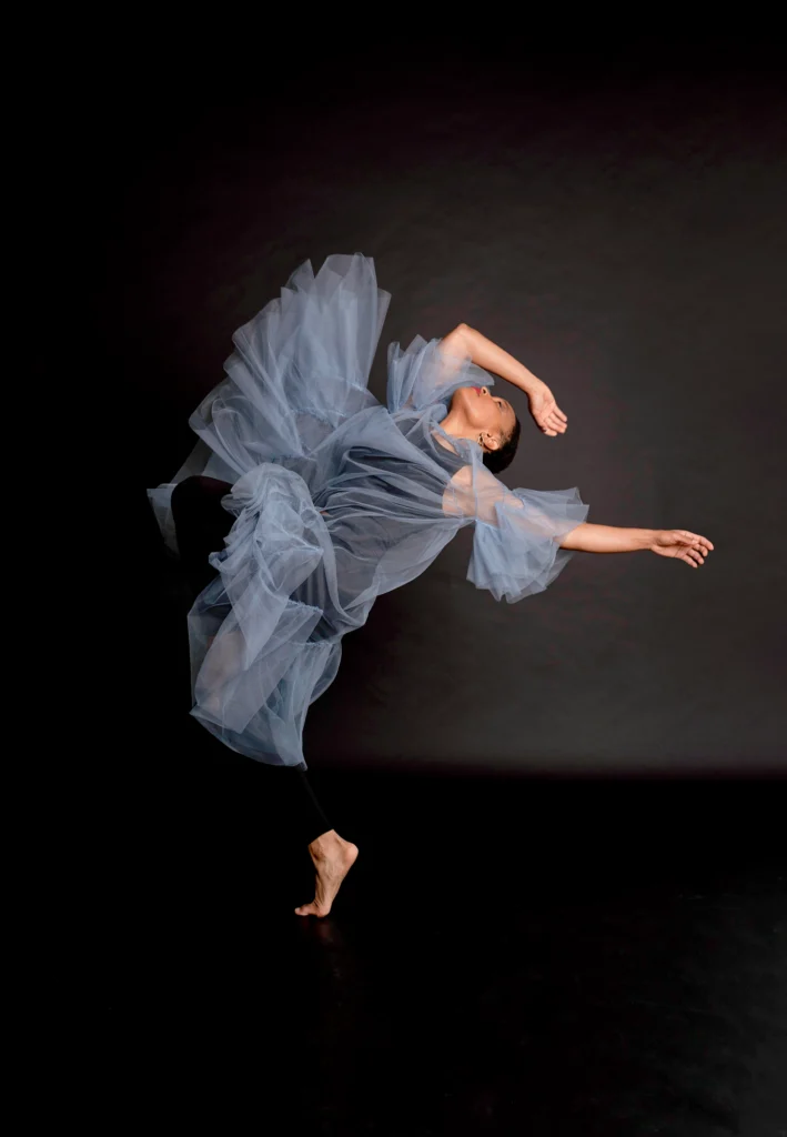 An African American woman on a black background dances wearing a blue flowing dress. She arches backward with one leg bent, one arm extended and the other arm bent above her head. Her eyes are closed.
