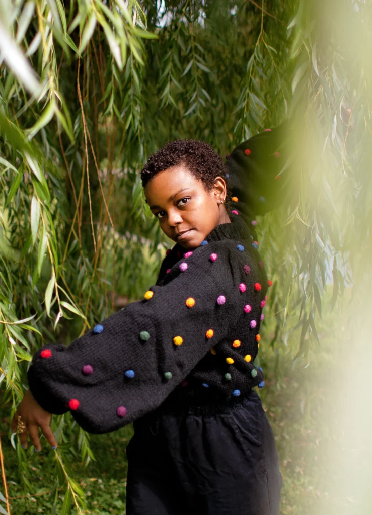 Laila J. Franklin gazes seriously at the camera from amidst trailing vines and greenery. Her hair is cropped close to her head; she wears a voluminous black sweater covered in multicolored puff balls. One arm curves down in front of her, the other twisting up behind her.