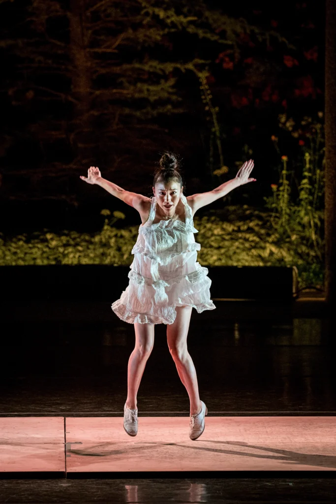 Naomi Funaki is caught mid pull-back, tap shoes hovering above the floor. Her arms fly behind her, but she gazes intensely forward. She is costumed in a grey-white puffy dress that matches her shoes. Her dark hair is piled in a bun atop her head. Greenery is visible beyond the stage.