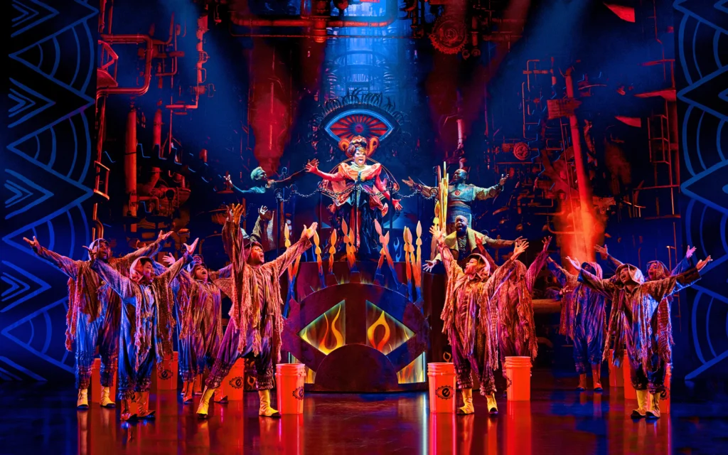 The stage is awash in reds and dark blues, evoking flame, as a dozen performers cluster and sing. Melody Betts stands atop a raised platform.