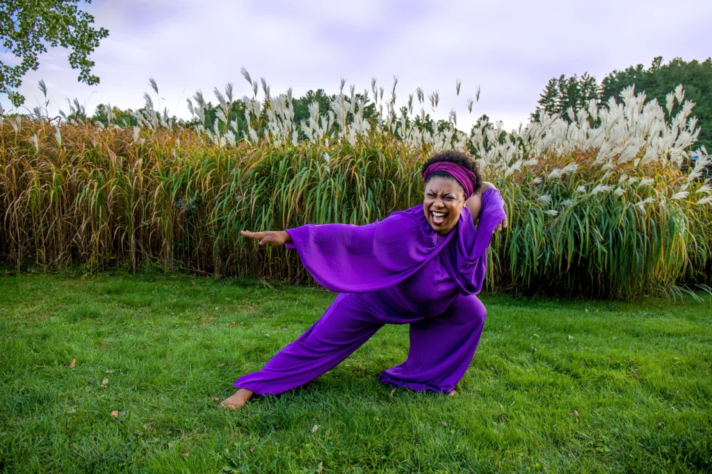 Sydnie Mosley, a Black woman wearing a flowy purple jumpsuit lunges back with her arms out. Her short black afro is held back by a purple scarf, her face shows a clear expression of joy. She is standing barefoot in front of the natural background of Ashfield, Massachusetts.