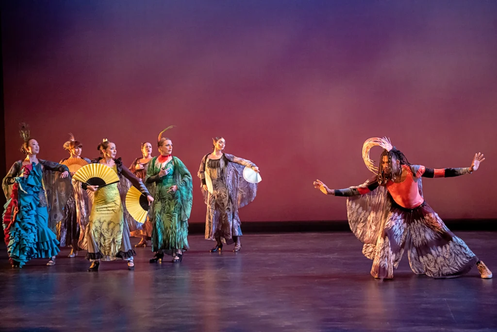 A half dozen colorfully dressed women flutter fans and look askance as Darrius Strong serenely flows through a low lunge. He is costume similarly in bright colors and patterns that evoke plumage, but wears sneakers instead of heels.