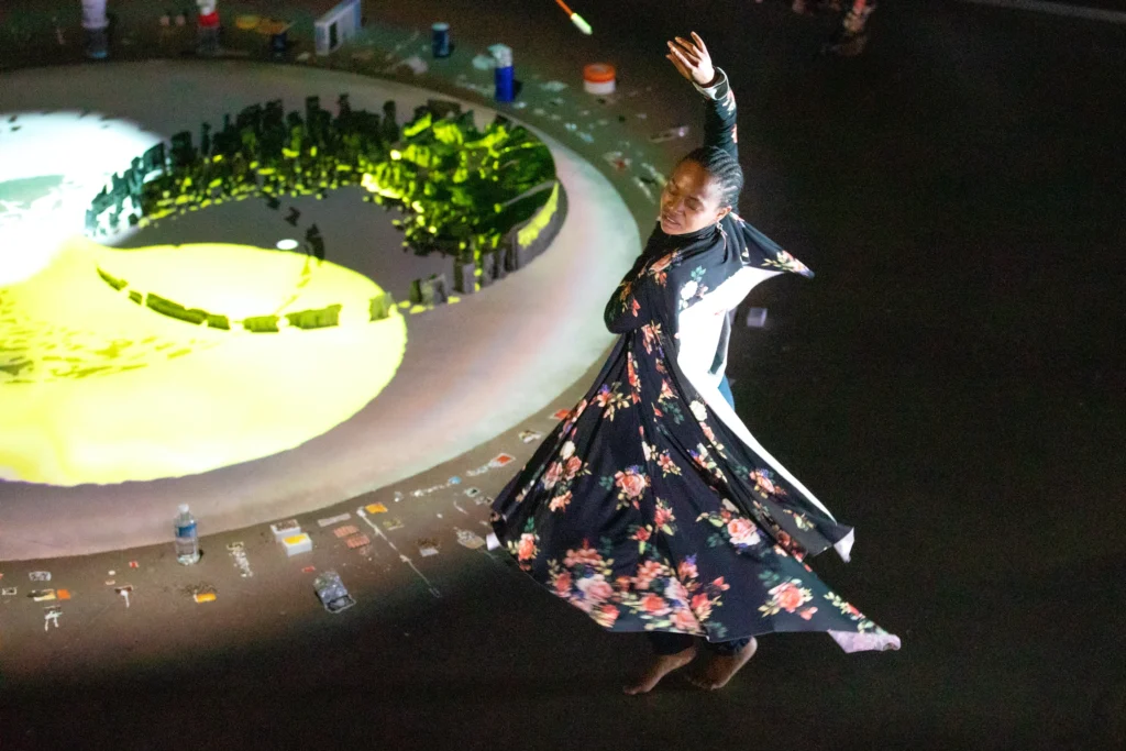 A flower-patterned black dress flares around a dancer as they turn, eyes closed, one arm elegantly overhead. They are at the outer edge of a circle on the ground formed by small objects and painted squares.