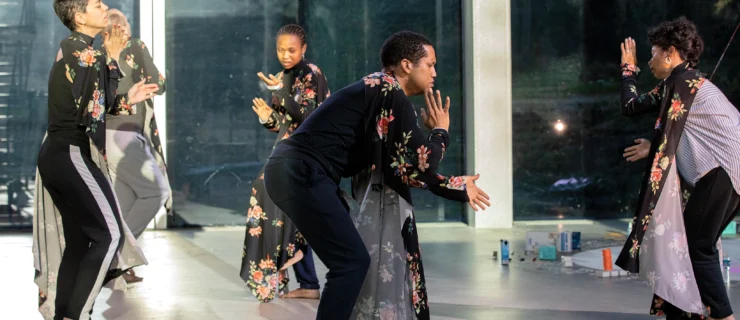 A loose circle of five dancers bend forward as they clap in unison, facing different directions. All are draped in black fabric with reddish pink flowers.