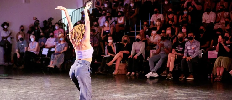 A dancer poses with their back to the camera, arms in a V overhead. A masked audience seated on risers watches.
